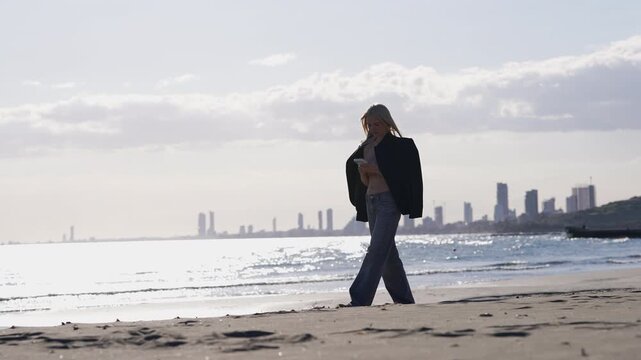 Young woman walking along the seashore with a smartphone in her hand. A modern city skyline is visible in the background against a sunny sky