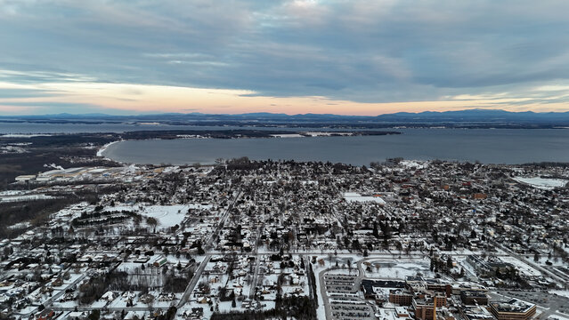Plattsburgh cityscape in clinton county, new york, with snow covered buildings and trees lining the shores of lake champlain at sunset, featuring distant mountains