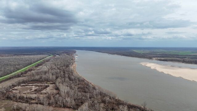 Expansive aerial view of the mississippi river from vidalia, louisiana, showing a wide waterway, bare winter trees along the banks, a levee, and a prominent sandbar under a cloudy sky