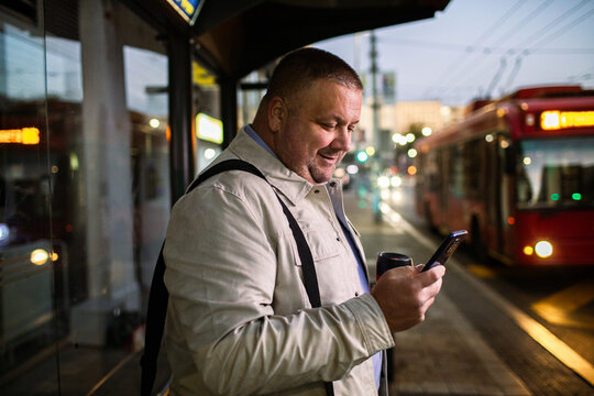 Smiling man using smartphone at city bus stop at dusk