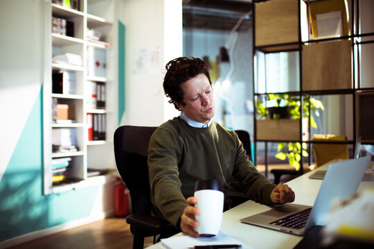 Tired office worker with coffee at laptop in modern office