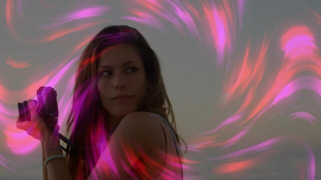 Woman standing shore holding camera looking left causing pink swirls circling head capturing sunset
