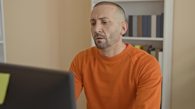Man in orange sweater covers face with hands at computer in building with bookshelf and notepad visible; stress burnout.
