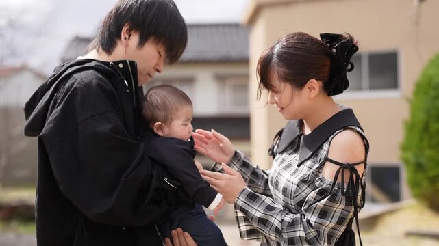 Man holds baby gently in park setting. Child smiles softly, eyes wide with wonder. Bright yellow tent stands behind bare trees. Distant pine tree adds depth to natural backdrop. Sunlight warms scene