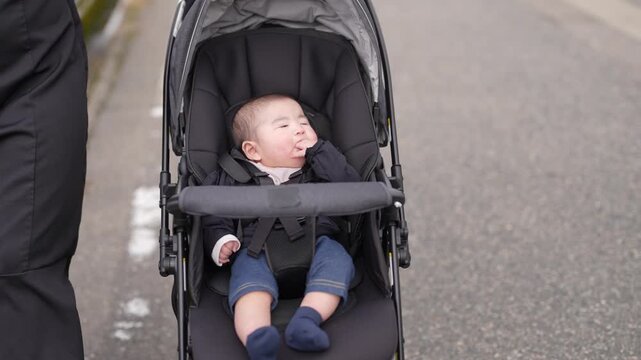 Soft light falls on a sleeping baby. Baby rests in a dark-colored stroller. Child's eyes are closed, face relaxed. Stroller moves slowly down a paved path. Parent's leg is visible beside the stroller