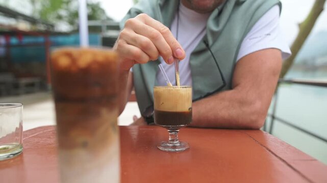 Chilly Iced Coffee Blurred With Stirring Spoon Outside In Fresh Air. Refreshing Outdoor Scene Featuring Small Espresso Glass With Creamy Foam And Blurred Iced Coffee In Background