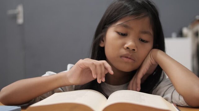Bored little Asian girl leaning on hand while reading a thick book, tired child feeling unmotivated or sleepy during homework, academic boredom and learning difficulties