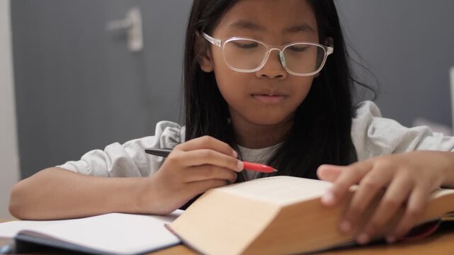 Diligent little Asian girl with glasses writing in notebook, focused child studying and reading book at home, early education and homeschooling
