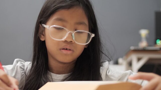 Diligent little Asian girl with glasses writing in notebook, focused child studying and reading book at home, early education and homeschooling