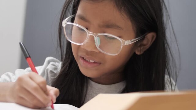 Stressed little Asian girl with glasses crying while studying, unhappy child having headache or difficulty with homework, academic pressure and learning disability