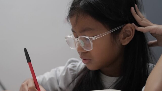 Stressed little Asian girl with glasses crying while studying, unhappy child having headache or difficulty with homework, academic pressure and learning disability