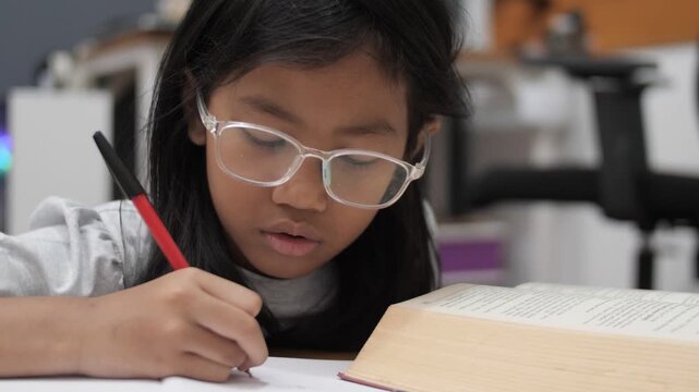 Stressed little Asian girl with glasses crying while studying, unhappy child having headache or difficulty with homework, academic pressure and learning disability