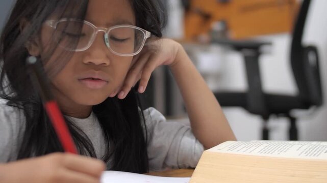 Stressed little Asian girl with glasses crying while studying, unhappy child having headache or difficulty with homework, academic pressure and learning disability