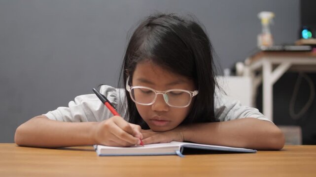 Diligent little Asian girl with glasses writing in notebook, focused child studying and reading book at home, early education and homeschooling