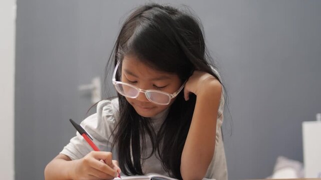 Diligent little Asian girl with glasses writing in notebook, focused child studying and reading book at home, early education and homeschooling