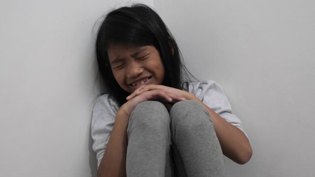 Sad little Asian girl crying in the corner of a room, lonely child sobbing and hiding in fear, concept of bullying, domestic problems