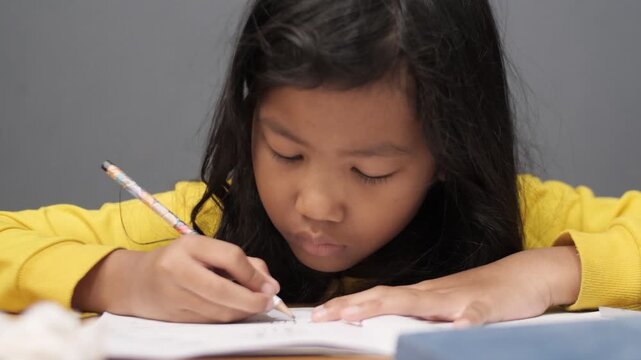 Diligent Asian little girl in yellow sweater writing on her notebook. Close up of focused elementary student doing homework or drawing. Childhood education, homeschooling