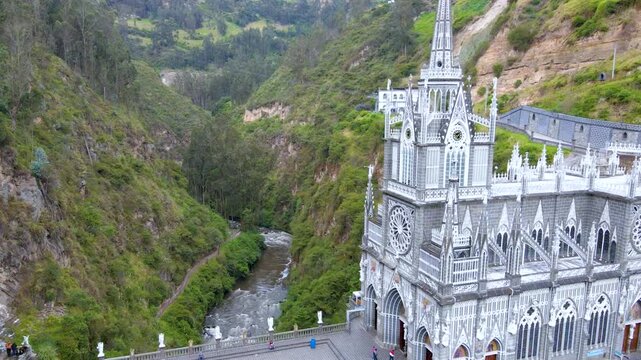 Drone view of Las Lajas Sanctuary perched on a stone bridge over a flowing river, surrounded by steep green cliffs and people walking on the plaza.