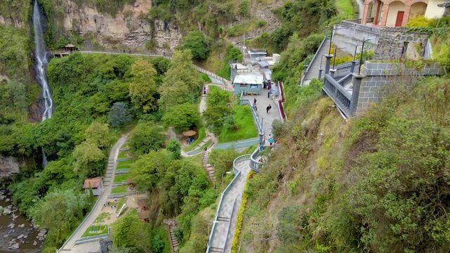 Drone view of Santuario de las Lajas in Ipiales showing a stone stairway and tall waterfall, visitors on terraces and a winding riverside path amid lush green cliffs.