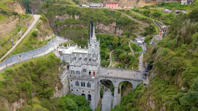 Aerial view of the gray Gothic sanctuary built into a steep canyon at Las Lajas, showing the arched bridge, flowing river below and people walking along the walkway.