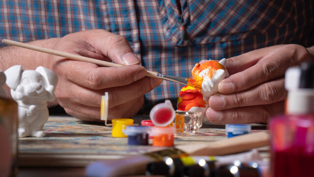 Close-up of a man's hands in a blue shirt holding a fine brush with orange paint, painting a small white ceramic figure