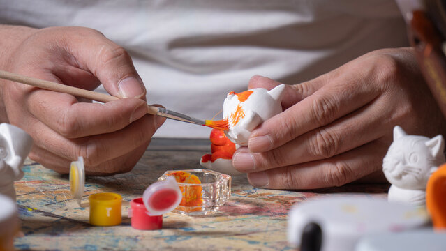 Close-up of a man's hands holding a fine brush with orange paint, painting a small white ceramic figure