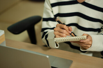 Fototapeta premium Closeup woman writing notes in small notebook beside laptop at desk, planning tasks