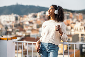 Fototapeta premium Relaxing woman drinking a cup of tea while listening to music with headphones on her balcony at home