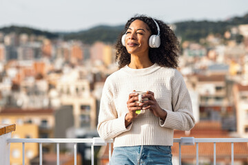 Fototapeta premium Relaxing woman drinking a cup of tea while listening to music with headphones on her balcony at home