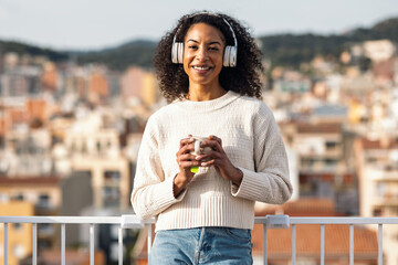Fototapeta premium Relaxing woman drinking a cup of tea while listening to music with headphones on her balcony at home