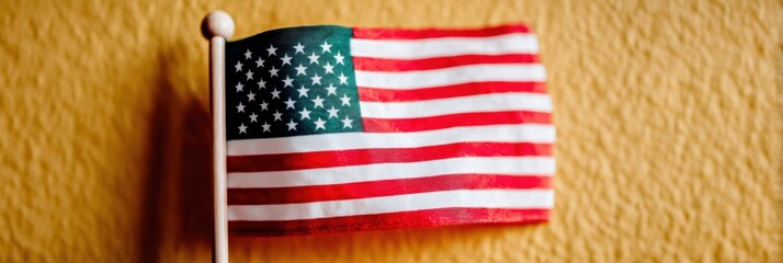 Obraz premium Small American flag on a pole stands against a yellow textured wall background for national themes, government education, and patriotic celebrations with copy space