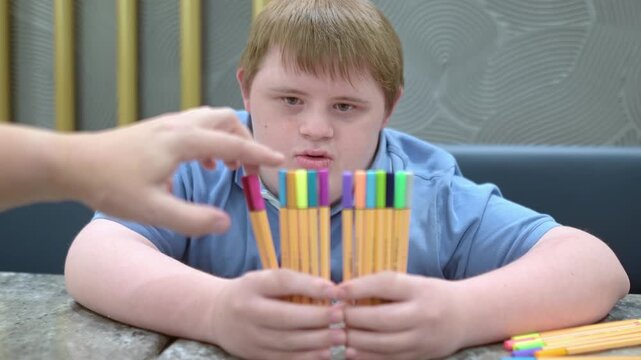 Teenager with Down syndrome having fun with markers. Boy plays game or counts pens after school. Integration of children and adults with disabilities. Lesson with tutor for children with special needs
