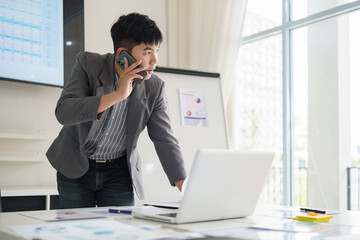 Naklejka premium Focused businessman talking on smartphone while reviewing data on laptop in modern office. Business communication and negotiation concept.