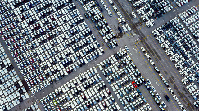 Aerial view of large car parking lot filled with new vehicles arranged in grid pattern at logistics distribution center during daytime, symbolizing automotive industry supply chain.