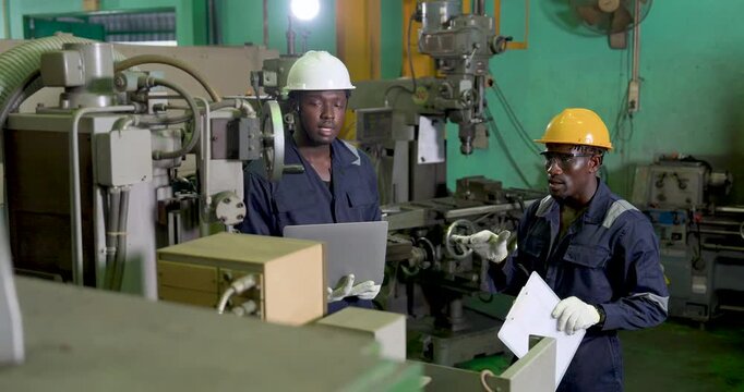 two african adult male engineers exchange production update beside machine area, using laptop and document in workshop, safety teamwork and workflow planning support decisions
