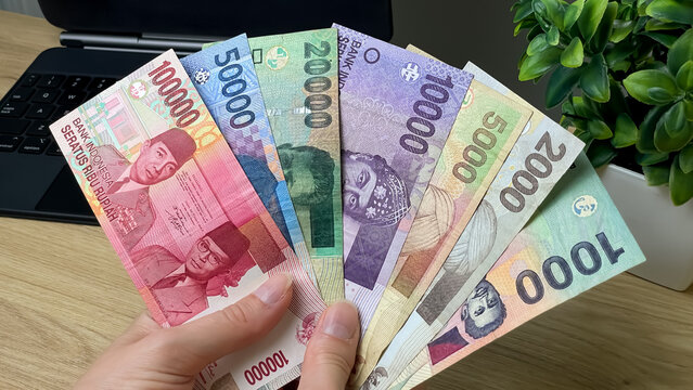 A hand holds a fan of Indonesian rupiah banknotes of various denominations against a background of a laptop and a plant. Suitable for topics such as finance, income, freelancing, and online work.