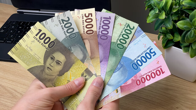 A close-up of a hand holding Indonesian rupiah banknotes against a laptop and plant. Conceptual ideas for finance, income, online work, and money management.