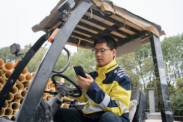 Worker operating a forklift while checking smartphone at outdoor construction site