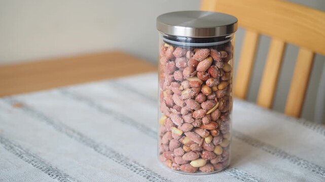 A close up view of a tall, clear glass jar filled with many roasted peanuts in shells on a white and striped tablecloth on a wooden table, with a chair in the background