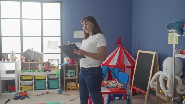 Middle age hispanic woman teacher holding clipboard and pen checking list in kindergarten building; calm focus.