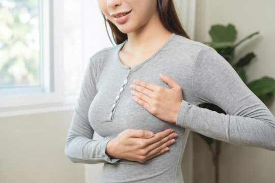 Health care breast cancer checking concept. Asian young woman, girl doing self check of breast cancer on gray t-shirt in room at home on white wall. How do I check breast cancer prevention awareness.