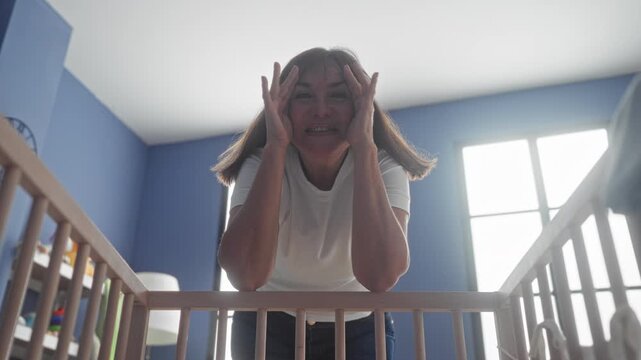 Woman leaning over crib covering her face with hands in a nursery inside a building; gentle playfulness.