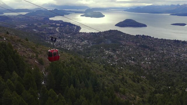 Right tracking aerial descent following a red cable car at Cerro Otto, Bariloche, Argentina, showing the town and Nahuel Huapi Lake on a partly cloudy day in 4K.