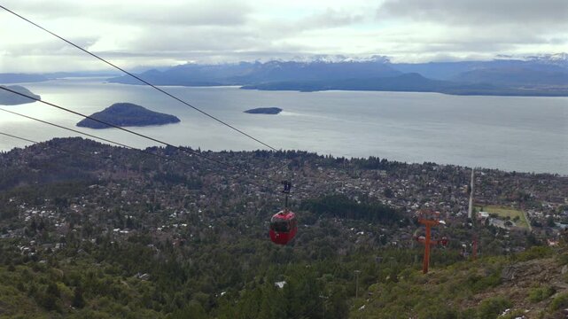 Red cable car descending at Cerro Otto in Bariloche, Argentina with Nahuel Huapi Lake and the city are visible in the background under clouds. Recorded at 4K. Static aerial shot.