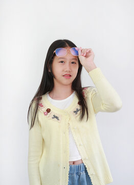 Trendy young Asian girl in pastel yellow cardigan against white background, pushing her stylish eyeglasses up onto her head while smiling at camera.