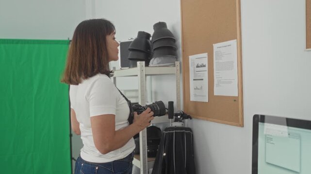 Woman photographer holds camera and inspects corkboard notes while standing by green screen and shelving in studio; concentration preparation.