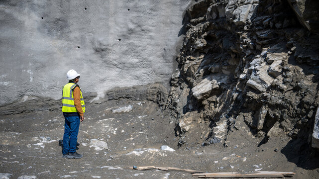 Geologist wearing safety helmet and reflective vest inspecting shotcrete rock face during excavation at a dam construction site. 