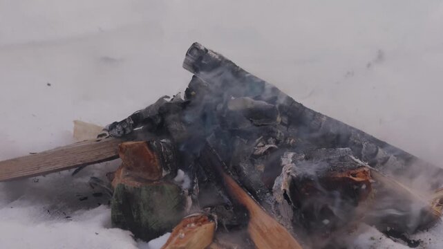 Close-up of smoking firewood remains inside a snow pit after the campfire has burned down. Charred logs and pale ash lie in the snow while smoke drifts upward. High quality 4k footage