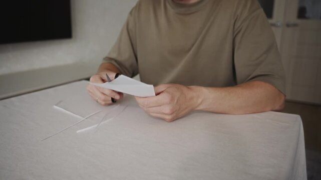 Man continues cutting a piece of white paper with scissors while sitting at a table. Paper strips accumulate on the table as the blades repeatedly close and open during the cutting process. 4k footage