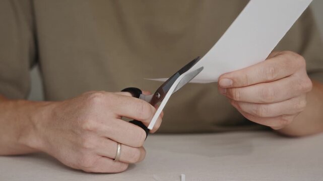 Hands using scissors to cut a piece of white paper on a table surface. Detailed view of simple craft or office task. High quality 4k footage